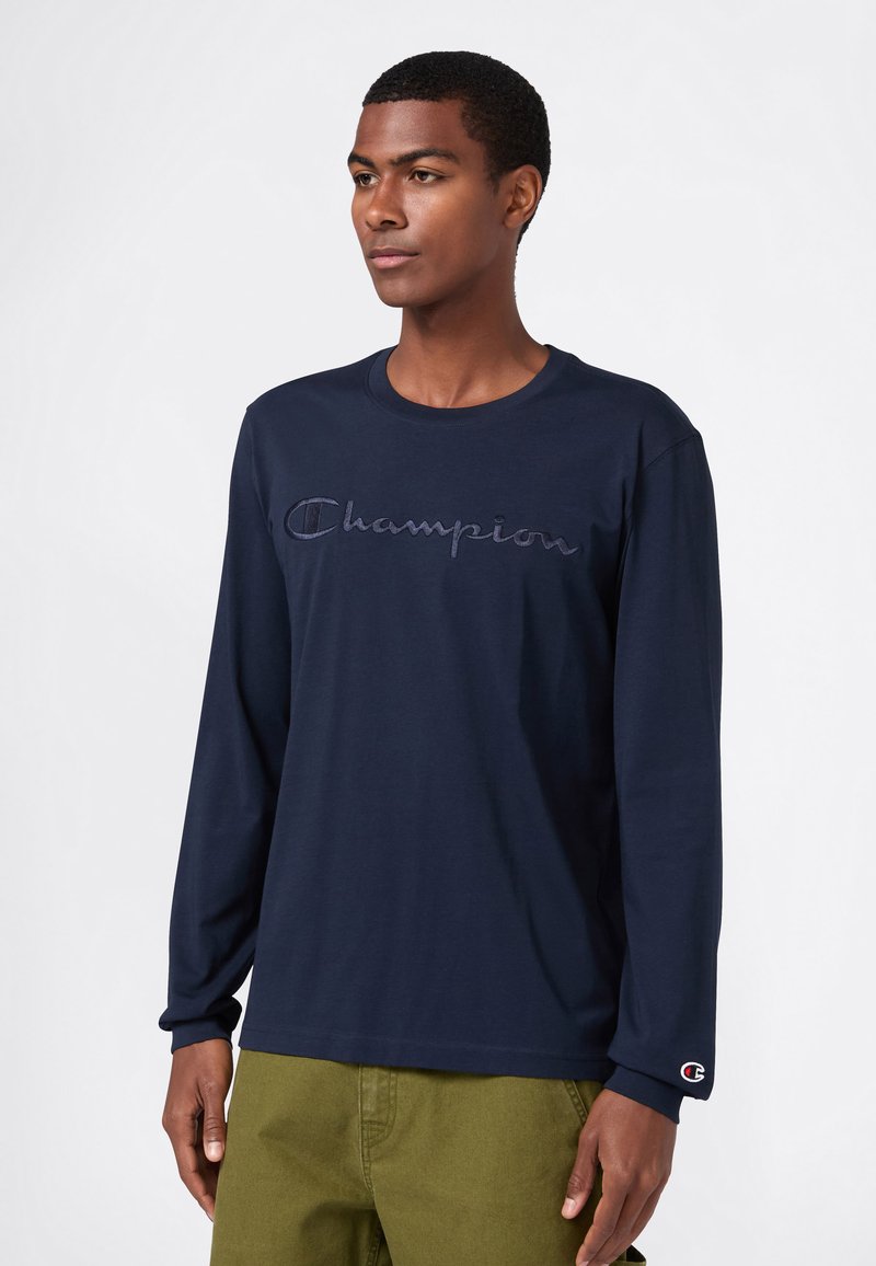 Junger Mann, der ein marineblaues Langarmhemd mit dem aufgestickten "Champion"-Logo auf der Brust und olivgrüne Hosen trägt, steht vor einem weißen Hintergrund.