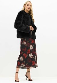 Chaqueta de piel sintética negra con mangas largas, combinada con una falda larga de estampado floral en rojos y blancos, complementada con sandalias de tiras negras.