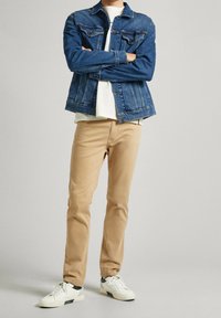 Chaqueta de mezclilla, azul claro, con dos bolsillos en el pecho, camiseta blanca debajo, pantalones beige y zapatillas blancas con acentos verdes.