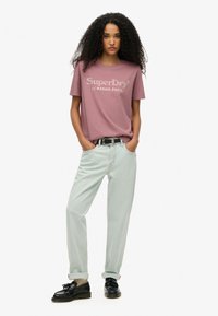 Mujer con cabello rizado que lleva una camiseta SuperDry color malva, jeans azul claro con puños, cinturón negro, calcetines beige y mocasines negros, de pie con las manos en los bolsillos.