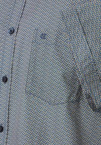 Short-sleeve, button-up shirt in blue fabric with a repeated yellow and white geometric pattern. Features a chest pocket and dark buttons.