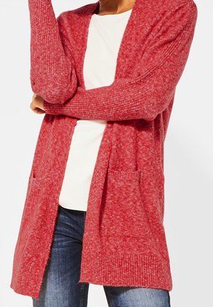 Person trägt eine lange, rote Strickjacke über einem weißen Hemd und blauen Jeans, die Arme vor der Brust verschränkt.