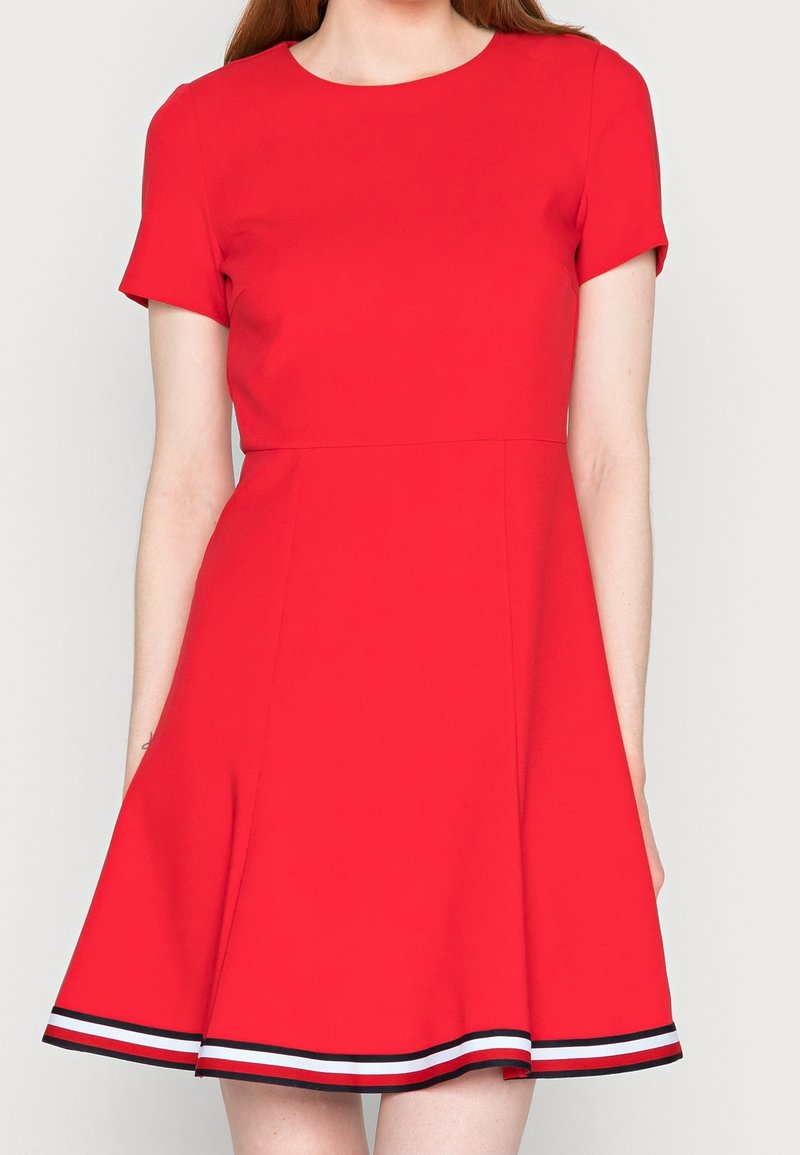 Femme portant une robe rouge à manches courtes avec une jupe évasée et une bordure rayée noire, blanche et rouge le long de l'ourlet.