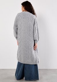 Dlouhý šedý pletený cardigan s texturovanými kabelkovými vzory a žebrovaným lemem. Rukávy jsou spadlé, což doplňuje široké džíny.
