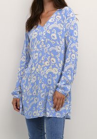 Tunique florale bleu clair avec des manches longues, un décolleté en V et une coupe décontractée. Le tissu a une texture avec des motifs floraux beiges.