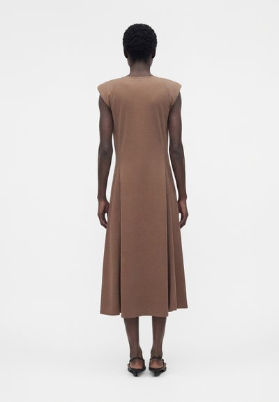 Άτομο που στέκεται με την πλάτη γυρισμένη, φοράει ένα καφέ μίντι φόρεμα χωρίς μανίκια με βάτες στους ώμους και μαύρα σανδάλια με χαμηλό τακούνι.