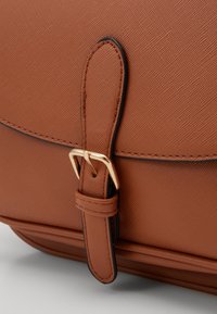 Ruskean teksturoidun nahkalaukun yksityiskohta, jossa on hihna, jossa on kultainen solki ja mustat ompeleen korostukset. Sileä pinta, jossa näkyvä rakeinen kuvio.