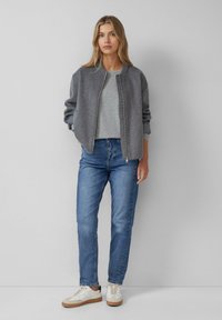 Veste bomber grise avec des poignets côtelés, portée sur un pull gris clair et un jean bleu. Des baskets blanches décontractées complètent la tenue.