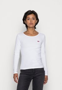 Witte katoenen t-shirt met lange mouwen, voorzien van een ronde hals en een klein rood logo op de linkerkant van de borst, gecombineerd met donkere jeans.