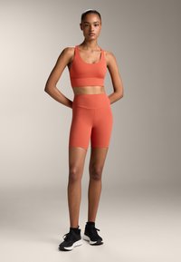 Piros-narancs sportmelltartó és hozzá illő, magas derekú, elasztikus anyagból készült rövidnadrág, fekete edzőcipővel párosítva. Simára szabott, testhezálló dizájn.