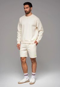 Felpa color crema e pantaloni corti realizzati in tessuto morbido, con un piccolo tag verde come accentuazione. Indossati con sneakers bianche e calze a righe.