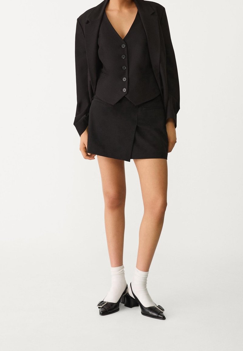Femme portant un blazer noir sur mesure, un gilet boutonné, une mini-jupe assortie, des chaussettes blanches et des chaussures à talons noires avec boucles sur fond blanc.