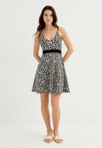 Vestido de patrón en blanco y negro con cuello en V, tirantes ajustables, cintura entallada y falda acampanada. El modelo lleva sandalias ligeras tipo slide.