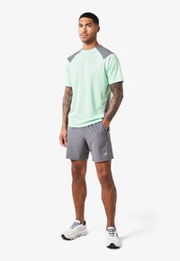 Camisa deportiva verde menta con acentos grises en los hombros, combinada con pantalones cortos grises. El modelo lleva zapatillas blancas, mostrando un look deportivo.