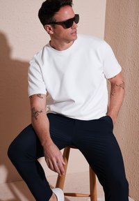 Camiseta blanca de manga corta hecha de tela suave, combinada con pantalones oscuros color navy. Sentado en un taburete de madera contra un fondo texturizado.