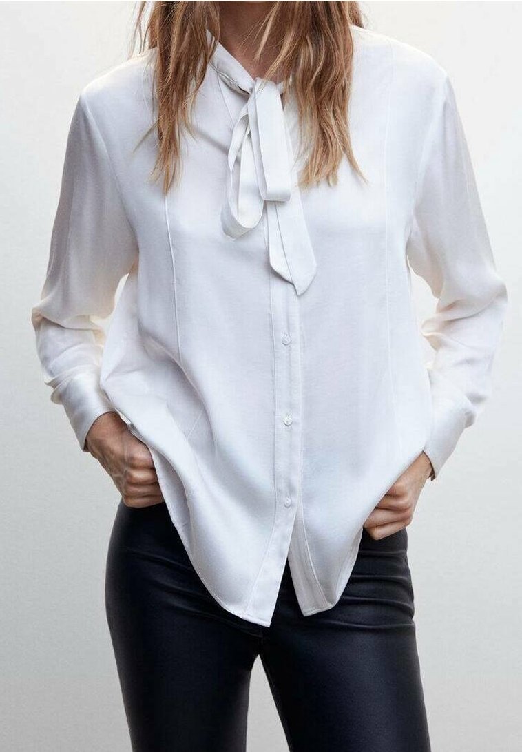 Femme portant un chemisier blanc à manches longues avec un nœud noué au col et un pantalon noir ajusté, les mains dans les poches.