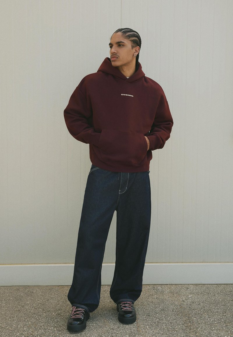 Giovane uomo con capelli intrecciati che indossa un hoodie bordeaux, jeans larghi scuri e sneaker nere in piedi contro un muro chiaro.
