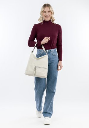 Femme souriante en train de marcher, portant un col roulé bordeaux, un jean bleu, des baskets blanches, et portant un grand sac à main beige à son bras.