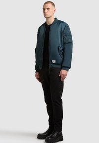 Man bär en mörk teal bomberjacka, svarta byxor och svarta stövlar, stående mot en enkel ljusgrå bakgrund.