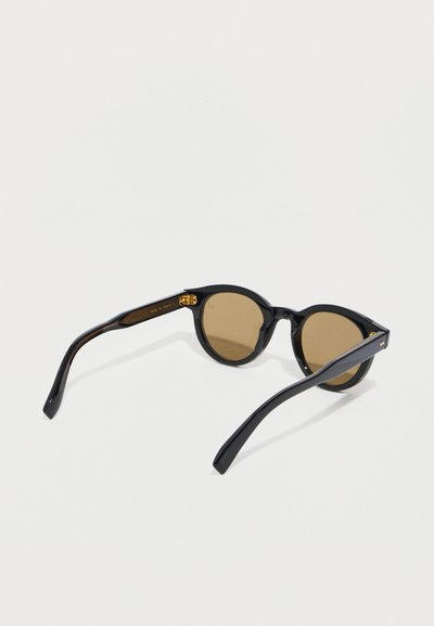 Lunettes de soleil à monture ronde noire avec des verres teintés marron et des détails de charnière dorés, posées sur une surface blanche.
