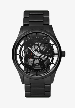 Sort analogur med et skeletdials, der har sølvfarvede time- og minutvisere, samt et metalrem og distinkte cirkulære former i designet.