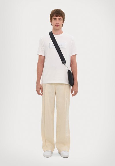 White cotton t-shirt with a black logo patch, beige wide-leg trousers, and white sneakers. Black crossbody bag with a textured strap.
