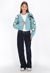 En ljusblå stickad cardigan med broderade vita blommor och texten "Smile", i kombination med mörka jeans och vita sneakers.