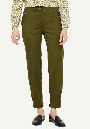 Pantalon vert olive sur mesure avec poches à rabat à l'avant et coutures latérales, associé à une chemise à manches longues à motif géométrique et des mocassins noirs.