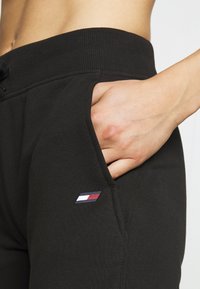 Musta collegehousut, joissa on resorivyötärö, sivutasku ja pieni logotagi, jossa on punaisia, valkoisia ja sinisiä yksityiskohtia vasemmalla puolella.