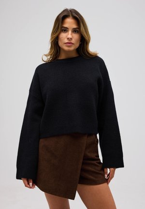 Joven con cabello ondulado suelto, vestida con un suéter negro de punto y una falda asimétrica marrón, de pie contra un fondo gris liso.