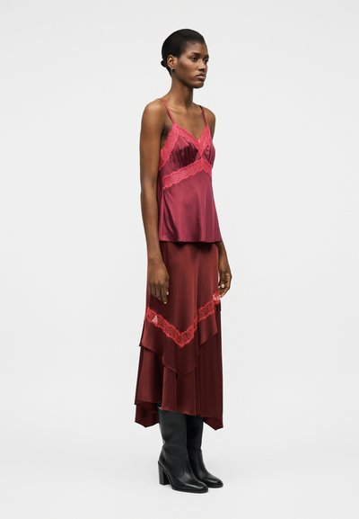 Γυναίκα που στέκεται φορώντας κόκκινη σατέν νυχτικό με δαντέλα, ταιριαστό στρωμένο φόρεμα και μαύρες μπότες με τακούνι μέχρι τη μέση της γάμπας σε λευκό φόντο.