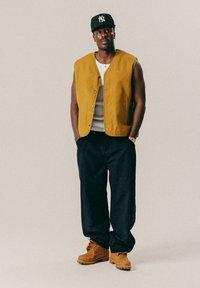 Mies, joka pukee mustaa lippistä, sinappikeltaista hihattomaa nappivaatetta, valkoista paitaa, mustia löysiä housuja ja ruskeita saappaita, seisoo kädet taskuissa.