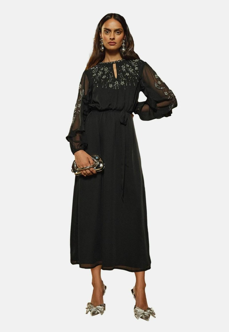 Rochie neagră cu mâneci lungi, cu un tiv brodat, mâneci transparente, talie legată și siluetă fluidă. Asortată cu o geantă metalică și pantofi cu toc în formă de fundă.