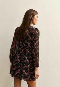 Vestido negro floral con mangas largas, tela transparente y cintura ajustada. Presenta patrones florales en rosa con puños con volantes y un dobladillo acampanado.