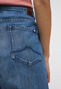 Vaqueros de denim en azul medio, que presentan un bolsillo trasero con un patrón de costura decorativa y una etiqueta de cuero en la cintura.
