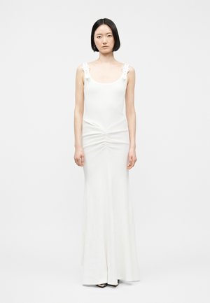 Kobieta o krótkich czarnych włosach, ubrana w białą, sięgającą podłogi suknię bez rękawów z kwiatowymi ramiączkami, stojąca na tle jednolitego jasnego tła.