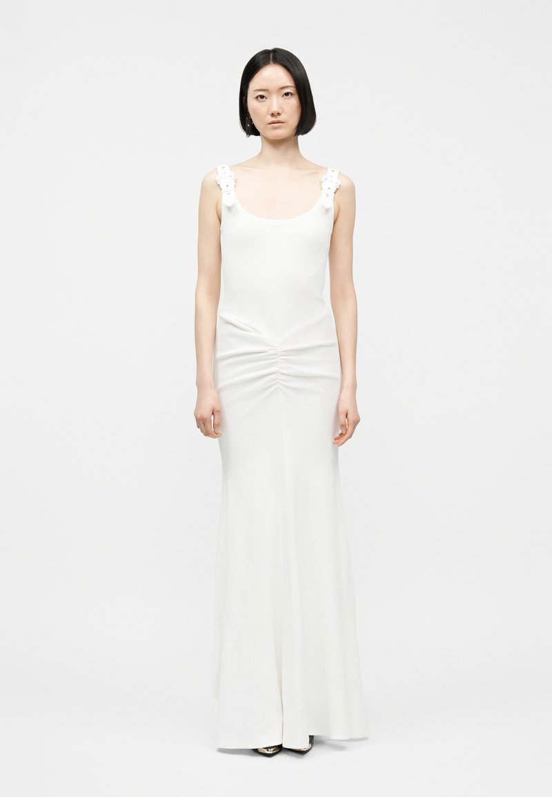 Γυναίκα με κοντά μαύρα μαλλιά που φοράει λευκό φόρεμα μέχρι το πάτωμα χωρίς μανίκια με λεπτομέρειες λουλουδιών στα λουριά, στέκεται μπροστά σε ένα απλό φωτεινό φόντο.