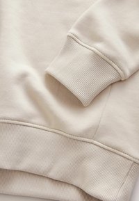 Tissu de sweat-shirt beige avec poignet côtelé. Détails des coutures visibles, texture lisse, design décontracté, avec une coupe ample et une apparence douce.