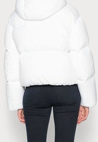 Biała kurtka puchowa o prostym kroju, błyszczącej fakturze, oversize'owym kapturze i elastycznym dole. Połączona z ciemnymi, dopasowanymi dżinsami.