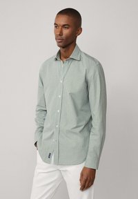 Camicia button-up verde a quadretti in tessuto leggero, con colletto aperto e maniche lunghe, abbinata a pantaloni bianchi.