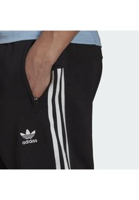 Pantalones deportivos negros con diseño de tres rayas en blanco, que cuentan con un bolsillo con cremallera y un logo bordado en el muslo izquierdo. Textura de tela suave.