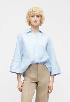 Jeune personne aux cheveux blonds courts et bouclés, portant un chemisier ample à rayures bleu clair et un pantalon beige ajusté, debout devant un fond uni.