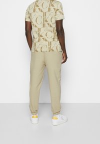 Beiges bomullströja och träningsoverallsbyxor med ett stort upprepat logotryck. Sneakers har vita och gula accenter. Bekväm och figurnära design.