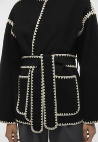 Schwarze Wolljacke mit weißen gewellten Nähten, weiten Ärmeln, einem taillierten Gürtel und zwei Fronttaschen mit abgerundeten Kanten.