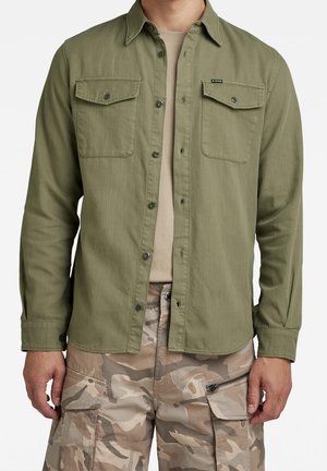 Homme portant une chemise boutonnée vert olive sur un t-shirt beige avec un pantalon cargo camouflage beige et marron.