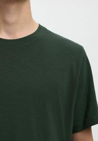 Camiseta de manga corta verde oscuro hecha de una tela suave y texturizada, con un cuello redondo y sutiles variaciones en el patrón del material.