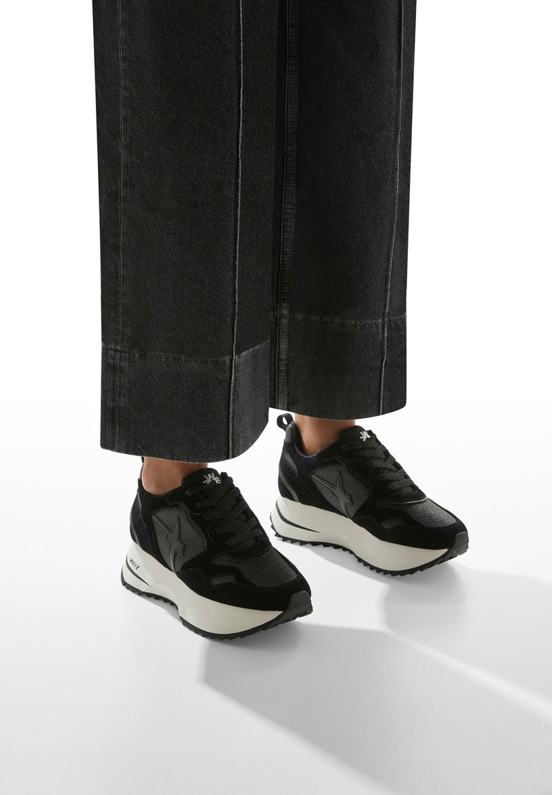 Baskets noires en matériau texturé, avec une semelle blanche et noire, et un logo en forme d'étoile sur le côté.