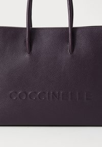 Purple leather tote bag with textured surface, featuring dual handles and embossed logo "COCCINELLE" on the front.