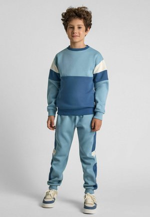 Sudadera de color azul claro con paneles en azul marino y blanco, combinada con joggers a juego que cuentan con acentos en los laterales. La tela parece suave y texturizada.