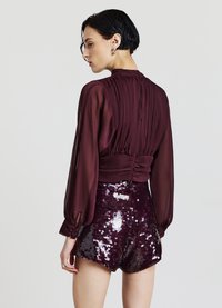 Blusa color bordeaux con arricciatura sul retro e maniche trasparenti abbinata a pantaloncini a vita alta ricoperti di paillettes viola lucide. Dettaglio testurizzato visibile.